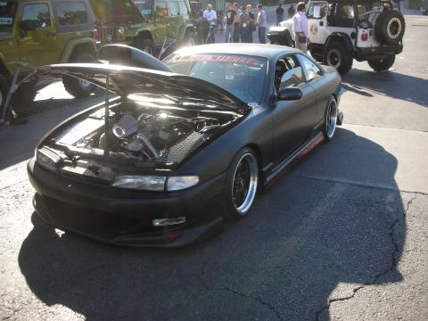 1998 Ford Crown Victoria - lowered matte-black sports coupe with an open hood and wide wheels sits at a car show beside Jeeps.