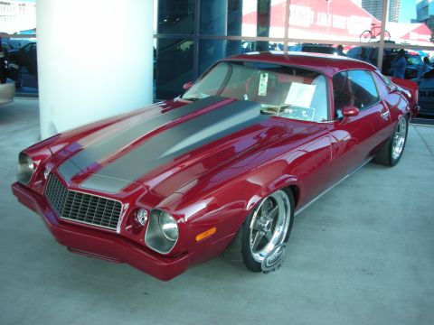 1998 Ford Crown Victoria - deep-red 1970s Camaro with a black hood stripe and polished five-spoke wheels sits at an indoor show.