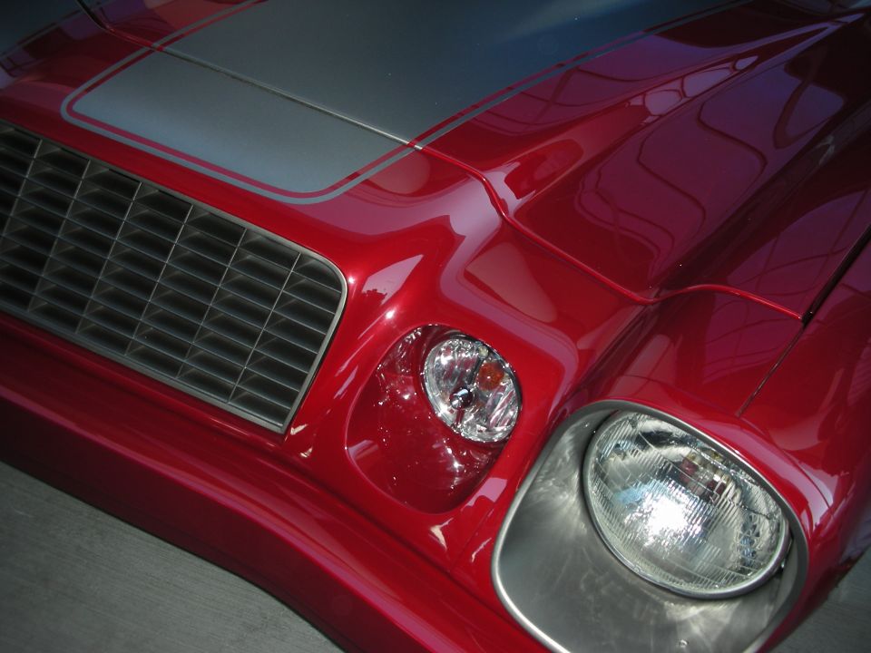 Front detail of a red Camaro showing headlight, grille and hood stripe