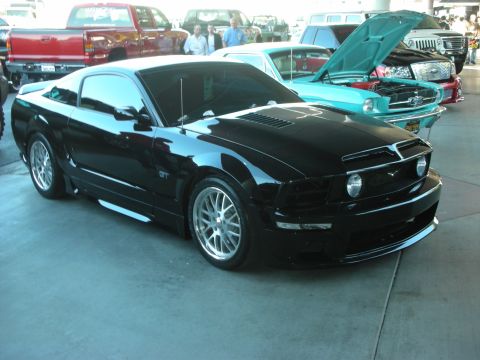 1998 Ford Crown Victoria - black Mustang GT with a custom body kit and polished wheels sits indoors beside a teal classic Mustang.