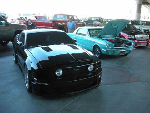 1998 Ford Crown Victoria - modified black Mustang GT sits beside a teal classic Mustang with its hood open at an indoor car show.