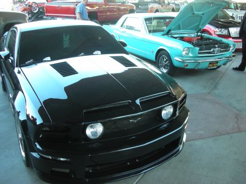1998 Ford Crown Victoria - Front view of a black Mustang GT with hood vents parked beside a teal vintage Mustang at an indoor show.