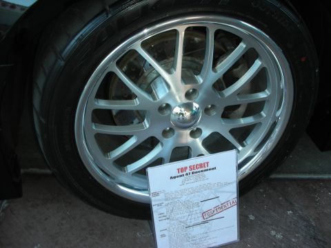 1998 Ford Crown Victoria - silver multi-spoke wheel and tire with a printed Top Secret build-spec sheet leaned against it.