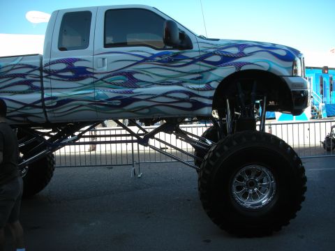 1998 Ford Crown Victoria - heavily lifted pickup with blue flame graphics shows its tall suspension links and oversized tire at a show.