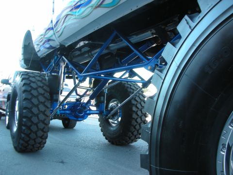 1998 Ford Crown Victoria - Low-angle shot under a lifted vehicle's flame-painted fender, showing a blue tubular suspension link beside an oversized 16.0 tire.