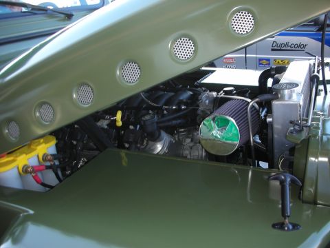 1998 Ford Crown Victoria - Engine compartment of an olive-green custom car with a louvered hood prop, green-and-chrome cone air filter and Duplicolor banner behind.