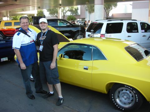 1998 Ford Crown Victoria - Two men stand smiling beside a yellow classic muscle car under a covered show area, with other vehicles parked nearby.