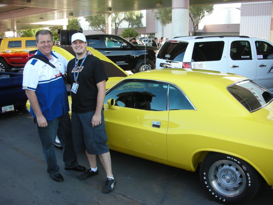 Two men posing beside a yellow muscle car at an outdoor car show