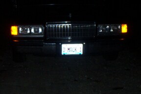1990 Mercury Grand Marquis - The front of a car is shown at night, illuminated by its headlights and license plate light.