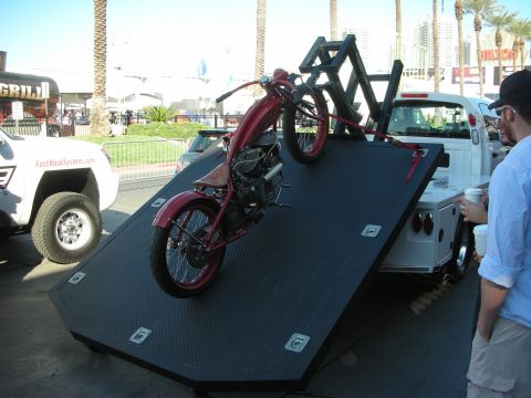 1998 Ford Crown Victoria - vintage red motorcycle sits strapped to the tilted flatbed of a tow truck at an outdoor event as a bystander looks on.