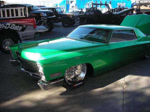 1998 Ford Crown Victoria - lowered green custom Cadillac coupe sits on chrome billet wheels at an outdoor show, with Jeeps and trucks in the background.