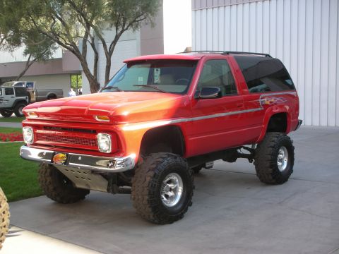 1998 Ford Crown Victoria - red lifted two-door Chevrolet SUV on large mud-terrain tires and chrome wheels parked on pavement beside a tree.