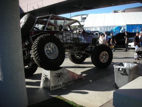 1998 Ford Crown Victoria - caged tube-chassis rock crawler perched on a display block on massive tires, with spectators walking past at an outdoor show.