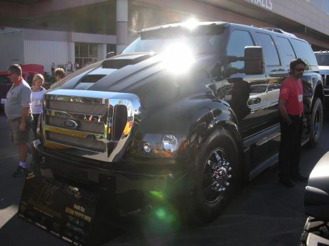 1998 Ford Crown Victoria - black customized medium-duty crew-cab truck with a large chrome Ford grille and cowl-hood, displayed indoors with onlookers and sun glare.