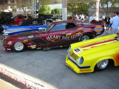 1998 Ford Crown Victoria - maroon Mustang-bodied drag funny car lettered HEART BREAKER beside a yellow flame-painted coupe under a covered show area.