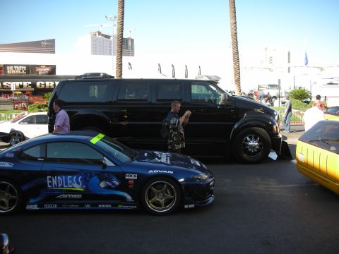 1998 Ford Crown Victoria - blue sponsor-liveried drift coupe parked beside a lowered black SUV with people and a city skyline behind.