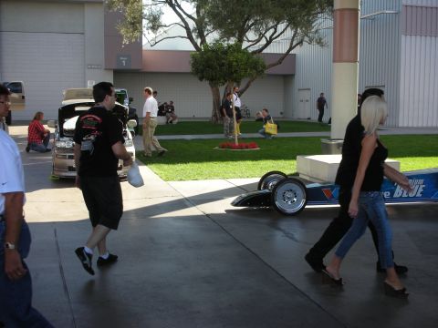 1998 Ford Crown Victoria - Show attendees walk past a long dragster chassis displayed on the pavement near a grassy courtyard.