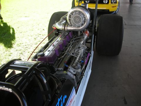 1998 Ford Crown Victoria - large polished turbocharger and intercooler piping mounted atop the exposed inline diesel engine of a dragster, viewed on grass.