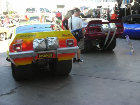 1998 Ford Crown Victoria - Rear view of a yellow-and-red flamed drag car with a PISAN01 plate beside a red car under a show tent with onlookers.