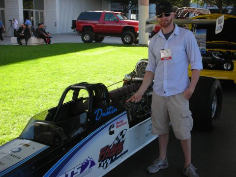 1998 Ford Crown Victoria - man in a ball cap stands beside a Cummins-powered dragster named Dustin, with a red SUV and lawn in the background.