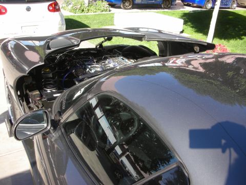 1998 Ford Crown Victoria - gray sports car with its rear clamshell open exposing the engine bay, parked on pavement with a white car behind.