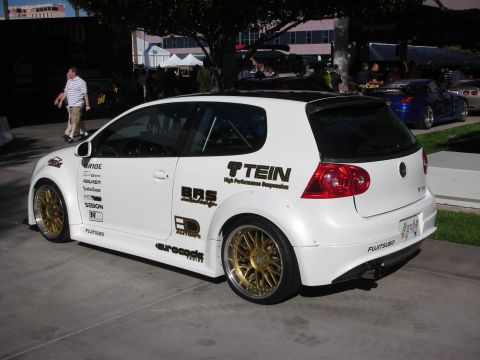 1998 Ford Crown Victoria - white Volkswagen Golf hatchback on gold mesh wheels covered in sponsor decals including TEIN and BBS, parked at a show.