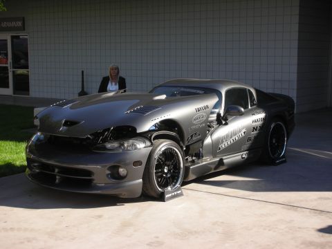 1998 Ford Crown Victoria - gunmetal Dodge Viper coupe with sponsor decals and wide rear drag tires displayed on a stand outside a building.