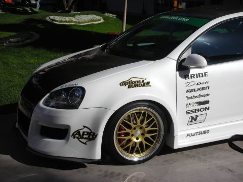 1998 Ford Crown Victoria - Close-up of the white Volkswagen Golf's front fender, projector headlight and gold mesh wheel, covered with sponsor decals.