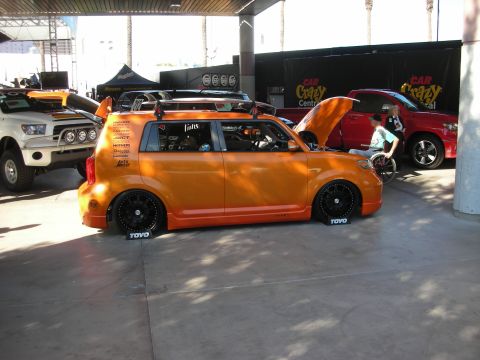 1998 Ford Crown Victoria - orange boxy Scion xB wagon, lowered on black wheels with Toyo tire markers, displayed under a covered car-show area.