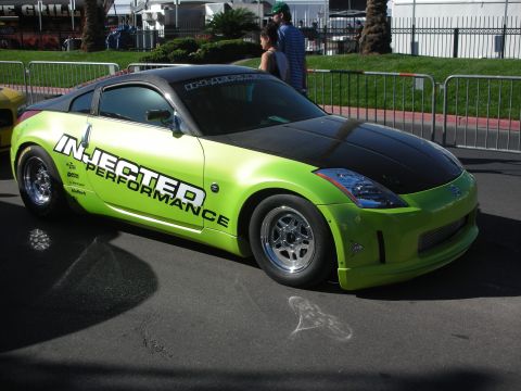 1998 Ford Crown Victoria - bright green Nissan 350Z drag car with a black hood and 'Injected Performance' side graphics parked on pavement near barricades.