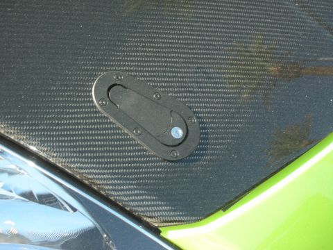 1998 Ford Crown Victoria - black flush hood-pin latch set into a carbon-fiber hood panel, with green bodywork visible at the lower edge.