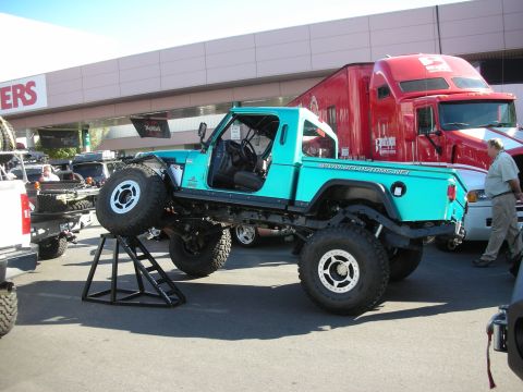 1998 Ford Crown Victoria - teal lifted Jeep CJ-style pickup with a removed front wheel sitting on a stand at an outdoor show, a red semi truck behind it.