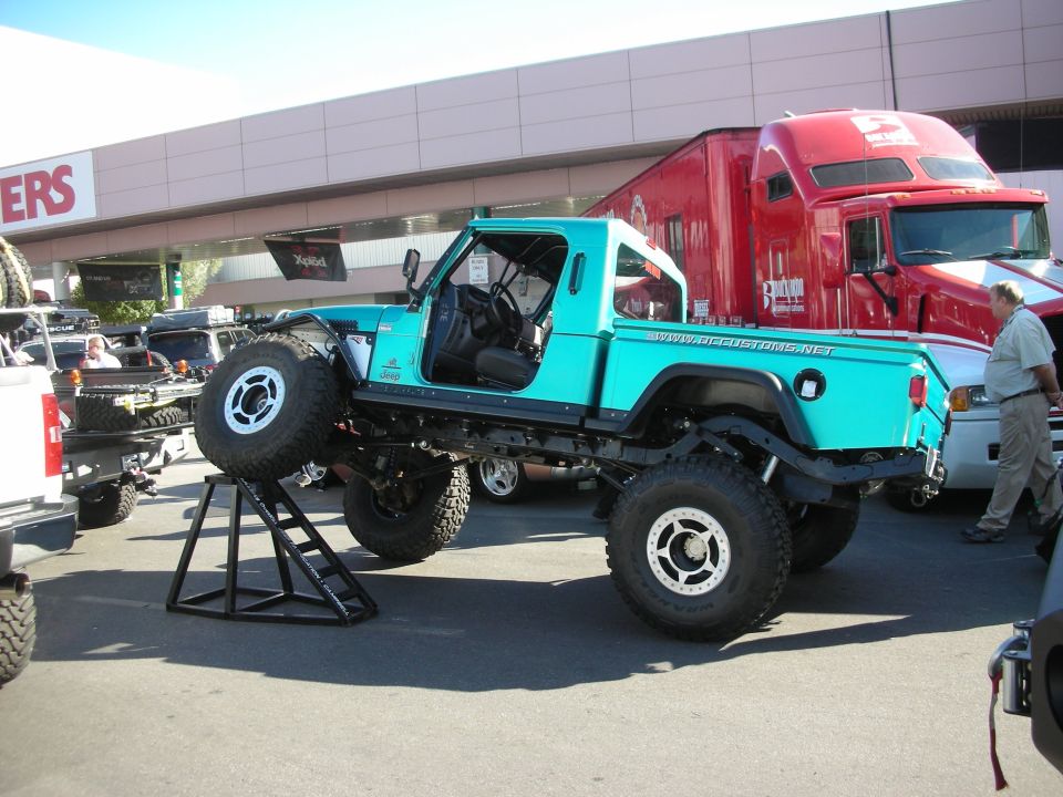 Teal lifted Jeep CJ pickup on a stand at an outdoor show with a red semi behind