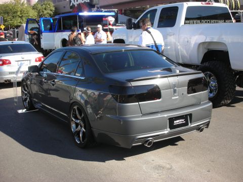 1998 Ford Crown Victoria - lowered gray custom sedan with a body kit and multi-spoke chrome wheels parked beside a lifted white pickup at an outdoor car show.