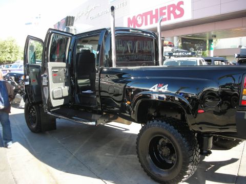 1998 Ford Crown Victoria - black Hummer H2 SUT dually pickup with a 4x4 badge and open rear door parked on a sidewalk in front of a Mothers booth.