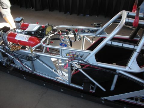 1998 Ford Crown Victoria - silver tube-frame dragster chassis fitted with a red fire-suppression bottle, wiring and roll-cage hoops on a black display stand.