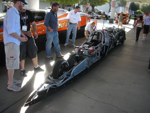 1998 Ford Crown Victoria - long black top-fuel style dragster with slick tires displayed on a sunny sidewalk while several spectators look on.