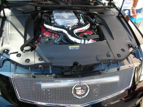 1998 Ford Crown Victoria - engine bay of a black Cadillac showing chrome intake piping over the V8 and a mesh grille bearing the Cadillac crest.