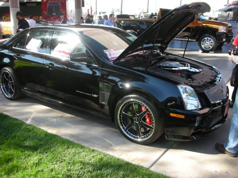1998 Ford Crown Victoria - black Cadillac performance sedan with its hood open, black multi-spoke wheels and red brake calipers displayed on grass at a show.
