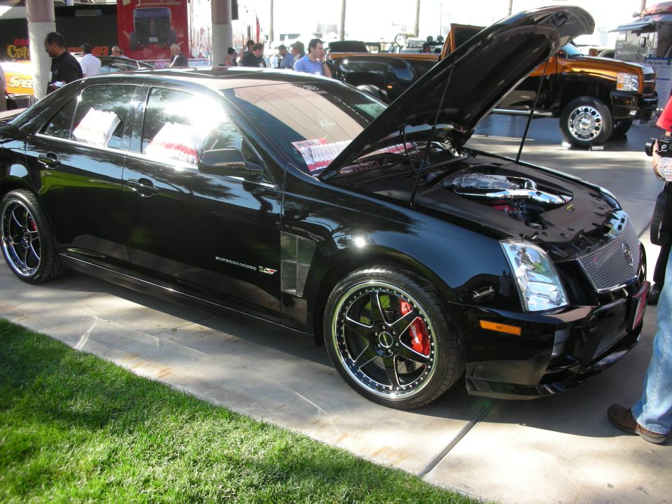 Black Cadillac sedan with open hood, black wheels and red brake calipers on grass at a show