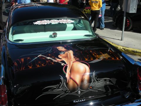 1998 Ford Crown Victoria - black car's trunk featuring an airbrushed pin-up woman mural with pinstriping, photographed at a crowded car show.