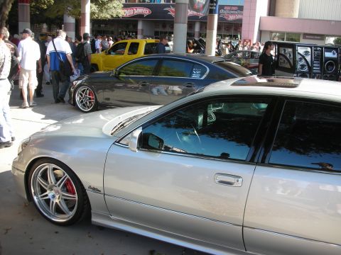 1998 Ford Crown Victoria - silver sedan and a gray sedan sit on polished aftermarket wheels at a busy outdoor automotive show.