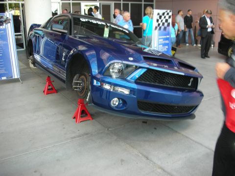 1998 Ford Crown Victoria - blue Shelby Mustang with white racing stripes rests on red jack stands, front wheels removed, at an indoor display.