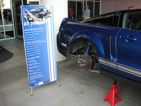 1998 Ford Crown Victoria - blue Mustang sits on a jack with brake rotor exposed next to a tall 'GT 700 KR' specifications banner.