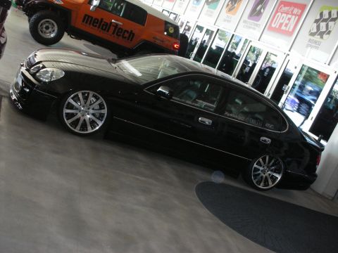1998 Ford Crown Victoria - black lowered luxury sedan on chrome wheels sits on a showroom floor in front of an orange off-road truck.