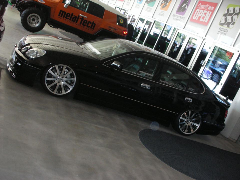 Black lowered luxury sedan on chrome wheels indoors near an orange off-road truck