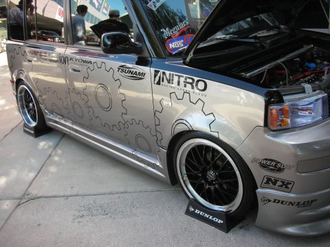 1998 Ford Crown Victoria - silver boxy wagon with mechanical gear graphics shows its open hood, black wheels, and sponsor decals at a show.