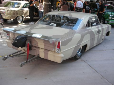 1998 Ford Crown Victoria - tan-and-brown camo drag car with rear wheelie bars and a Marines decal sits among show cars outdoors.