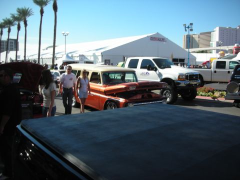 1998 Ford Crown Victoria - orange vintage pickup and a lifted white Ford Super Duty parked among crowds at an outdoor car show with palm trees and tents.