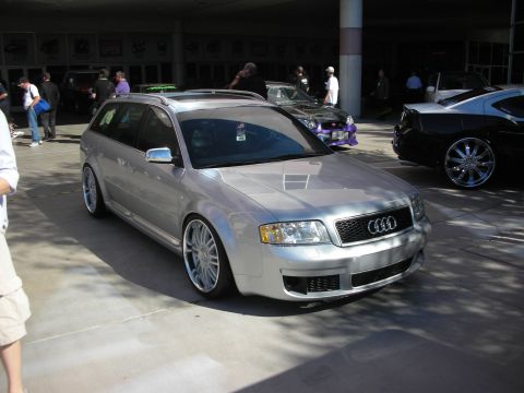 1998 Ford Crown Victoria - lowered silver Audi wagon with the four-ring badge and polished wheels sits indoors at a car show among other cars.